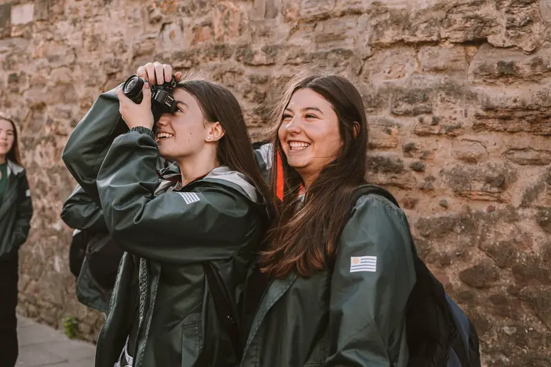 Dos viajeras riendo mientras sacan fotos junto a un muro de piedra